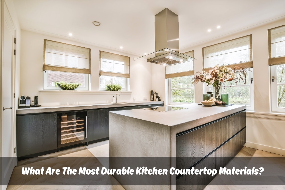 Modern kitchen featuring large soapstone island as centerpiece. Island showcases fruits and flowers in vase. Text overlay: "What Are The Most Durable Kitchen Countertop Materials?"