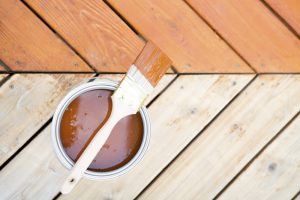 Open can of timber decking stain with brush on wooden deck, showing a comparison between stained and unstained timber decking.